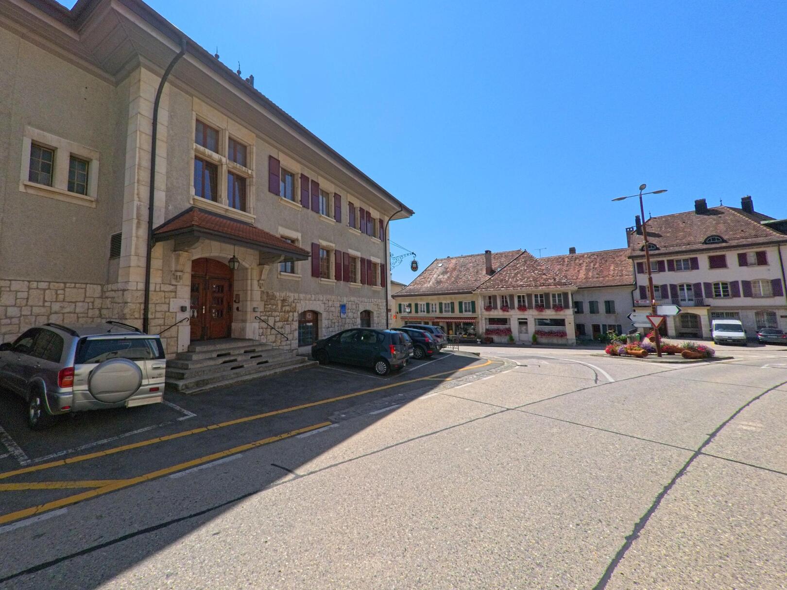 Im Zentrum von Bière (1145, Kanton Waadt) stehen historische Gebaeude mit roten Fensterlaeden und ein kleiner Kreisverkehr mit Blumenschmuck.