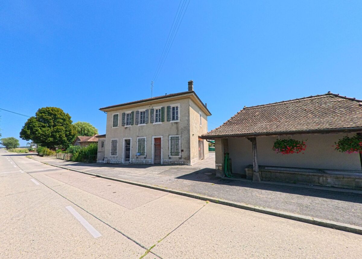 Ein altes Wohnhaus mit gruenen Fensterlaeden und ein ueberdachter Brunnen stehen in Bière (1145, Kanton Waadt) an einer breiten Strasse.