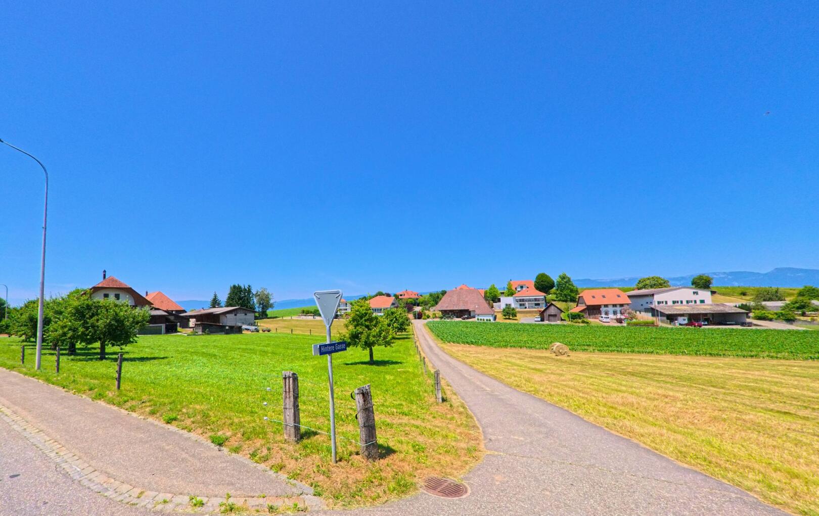 In Biezwil (4585, Kanton Solothurn) führt die Hintere Gasse als schmaler Asphaltweg durch eine offene Landschaft mit Bauernhäusern und Feldern bis zum Dorfkern.