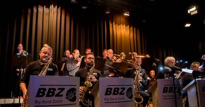 In Kloten ([ORT] Kloten, [PLZ] 8302, Kanton [KANTON] Zürich) spielt eine Big Band auf einer beleuchteten Bühne, waehrend mehrere Musiker mit Saxophonen und Blechblasinstrumenten gemeinsam musizieren und vor Notenstaendern sitzen.