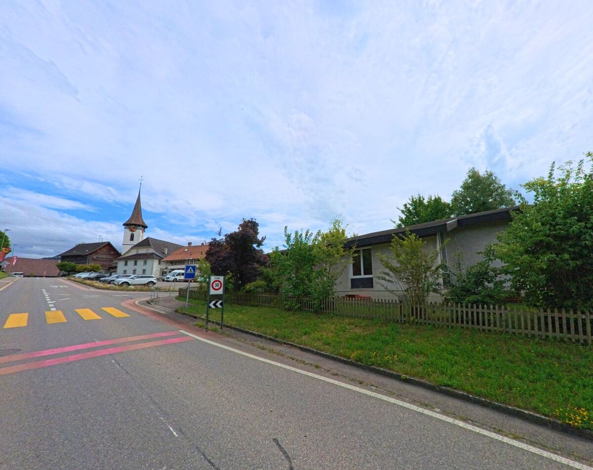In Biglen (3507, Kanton Bern) führt eine Hauptstrasse mit Zebrastreifen an einem flachen Gebäude vorbei direkt auf die Kirche mit ihrem spitzen Turm zu.