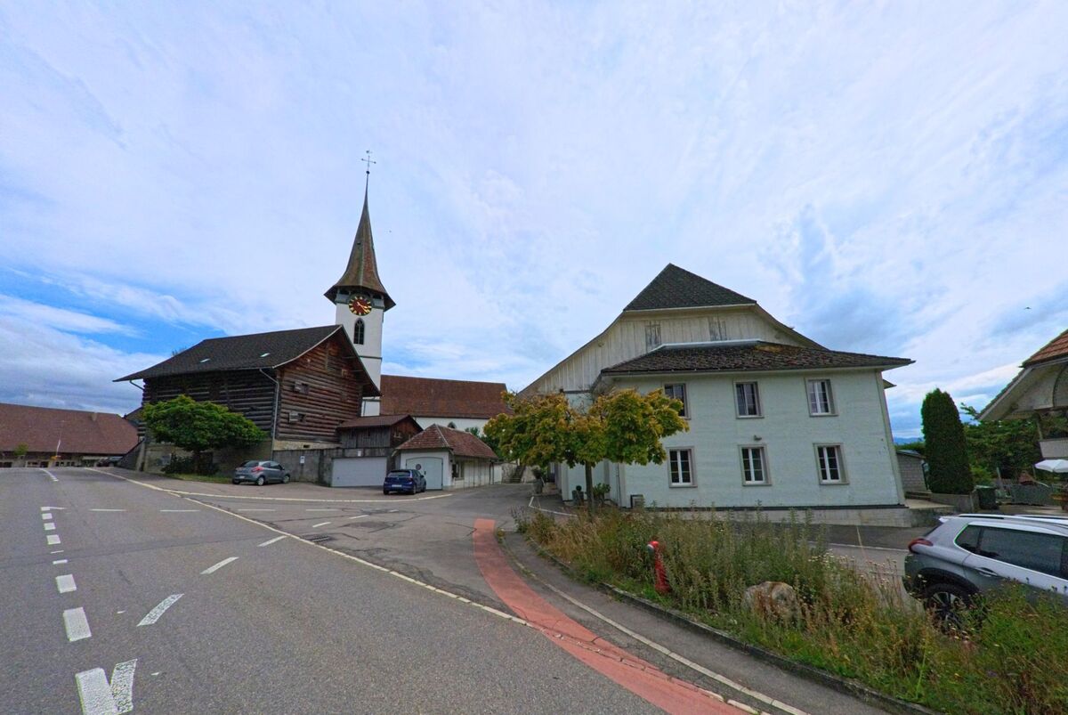 In Biglen (3507, Kanton Bern) steht die Kirche mit markantem Turm und Ziffernblatt an einer leicht ansteigenden Strasse, flankiert von traditionellen Gebäuden.