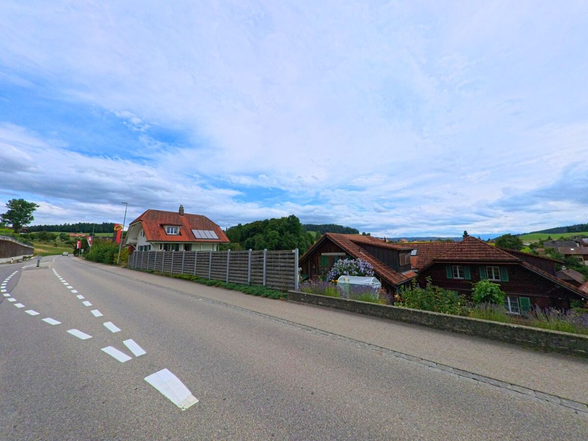 Am südlichen Ortsrand von Biglen (3507, Kanton Bern) verläuft eine Landstrasse oberhalb traditioneller Wohnhäuser mit Blick auf die umliegende Hügellandschaft.