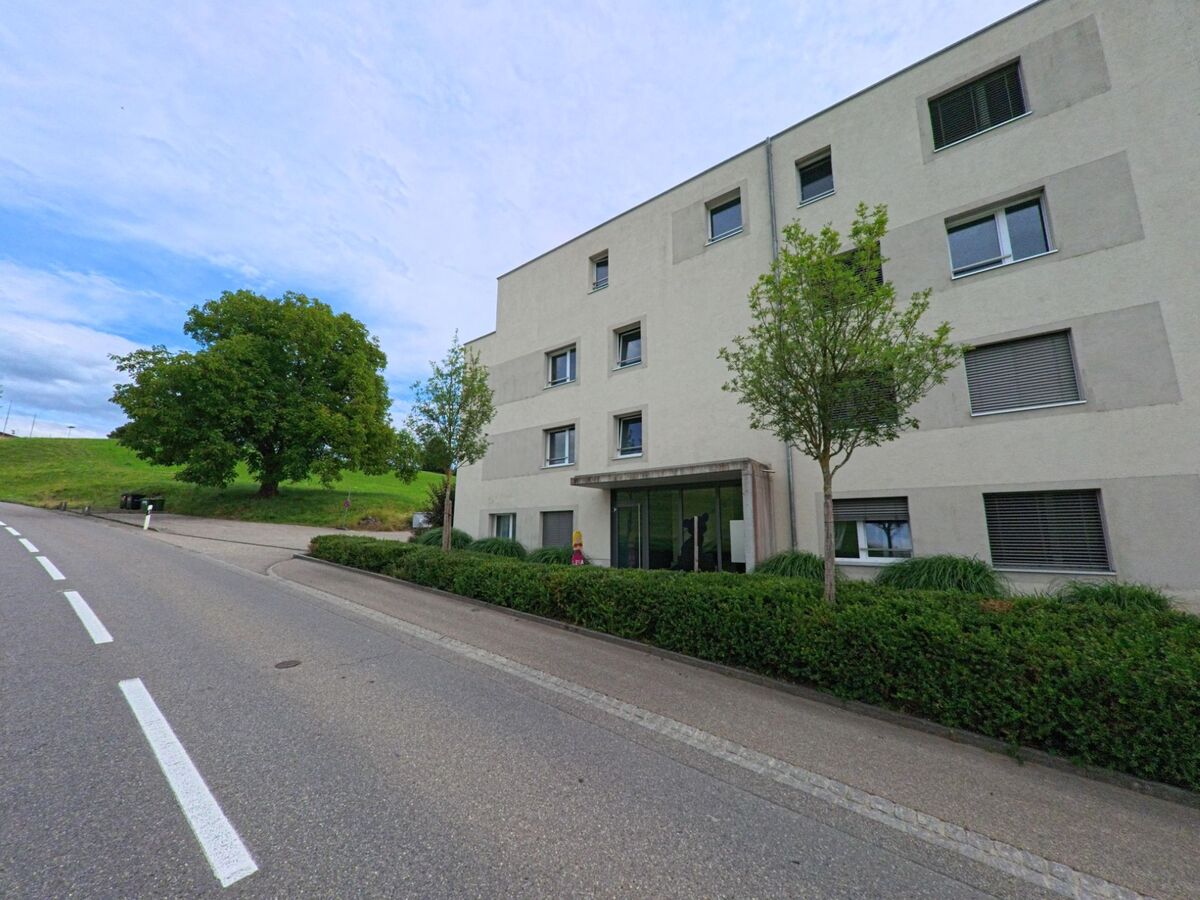 Ein modernes Wohngebäude mit klarer Fassadengestaltung steht in Biglen (3507, Kanton Bern) an einer leicht ansteigenden Strasse, umgeben von gepflegten Grünflächen.