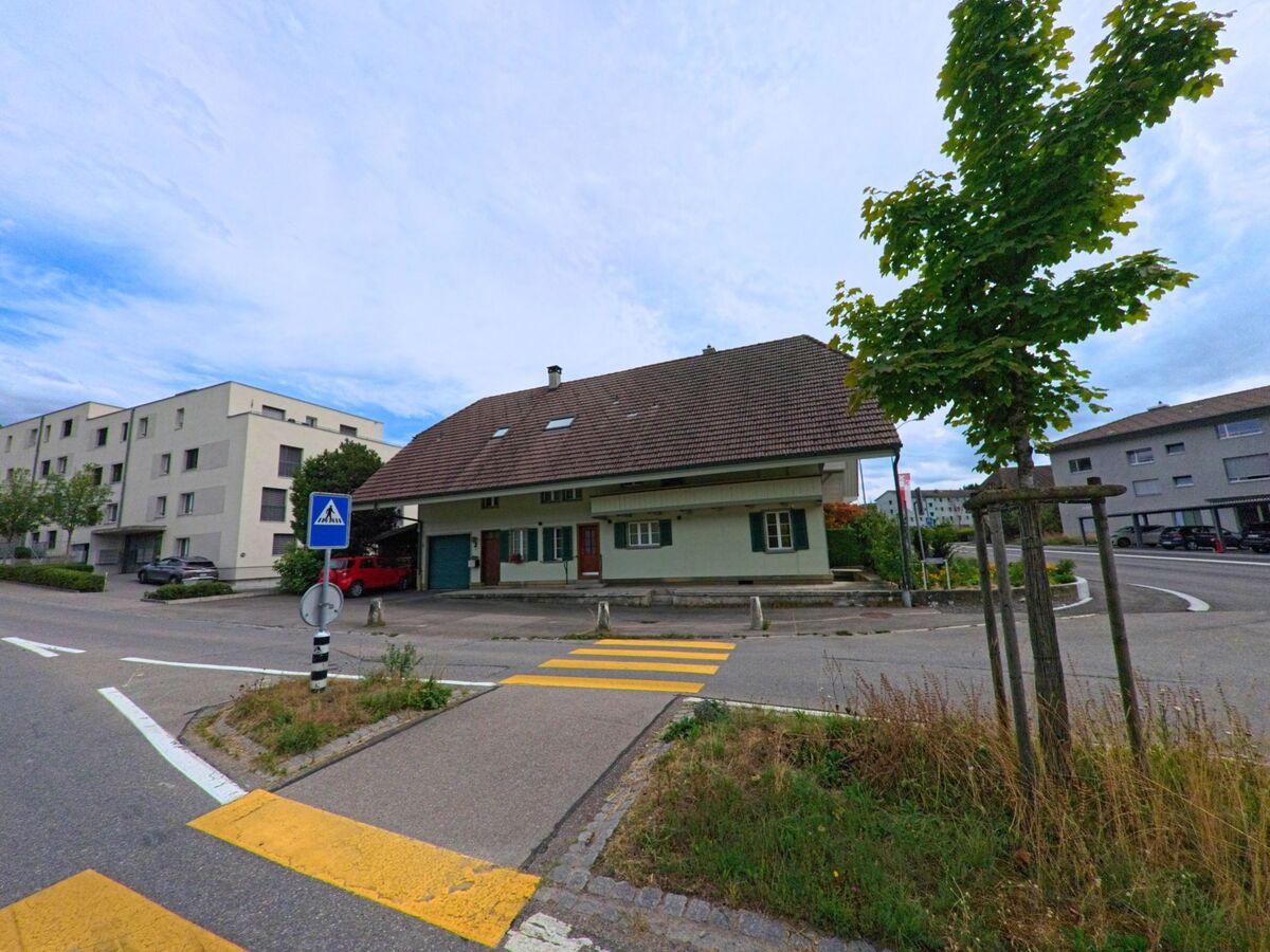  In Biglen (3507, Kanton Bern) steht ein traditionelles Wohnhaus mit grossem Dachfirst an einer Kreuzung, umgeben von modernen Mehrfamilienhäusern und einem Fussgängerstreifen.