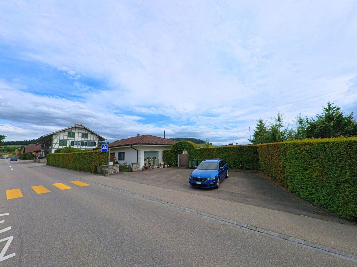 In Biglen (3507, Kanton Bern) verläuft eine Strasse mit Zebrastreifen an Einfamilienhäusern vorbei, gesäumt von gepflegten Hecken und einem kleinen Parkplatz.