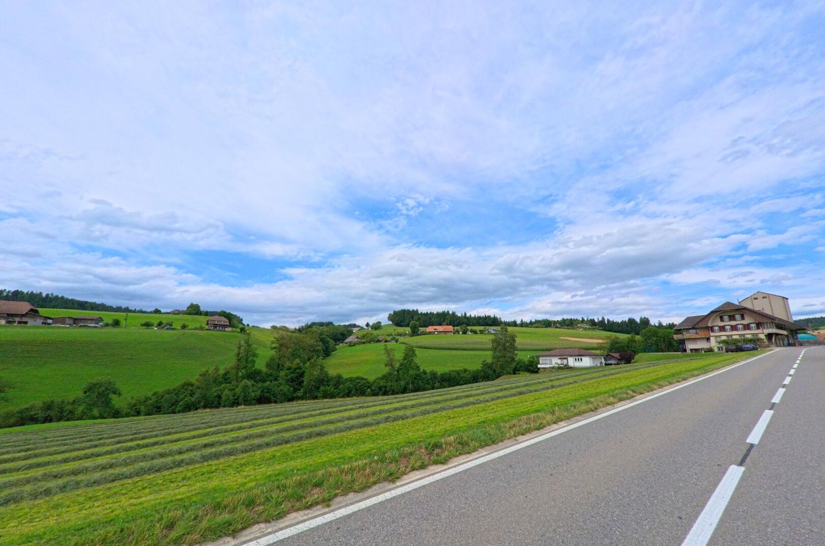 Eine Landstrasse führt durch die hügelige Landschaft von Biglen (3508, Kanton Bern) mit grünen Feldern und Bauernhäusern unter leicht bewölktem Himmel.