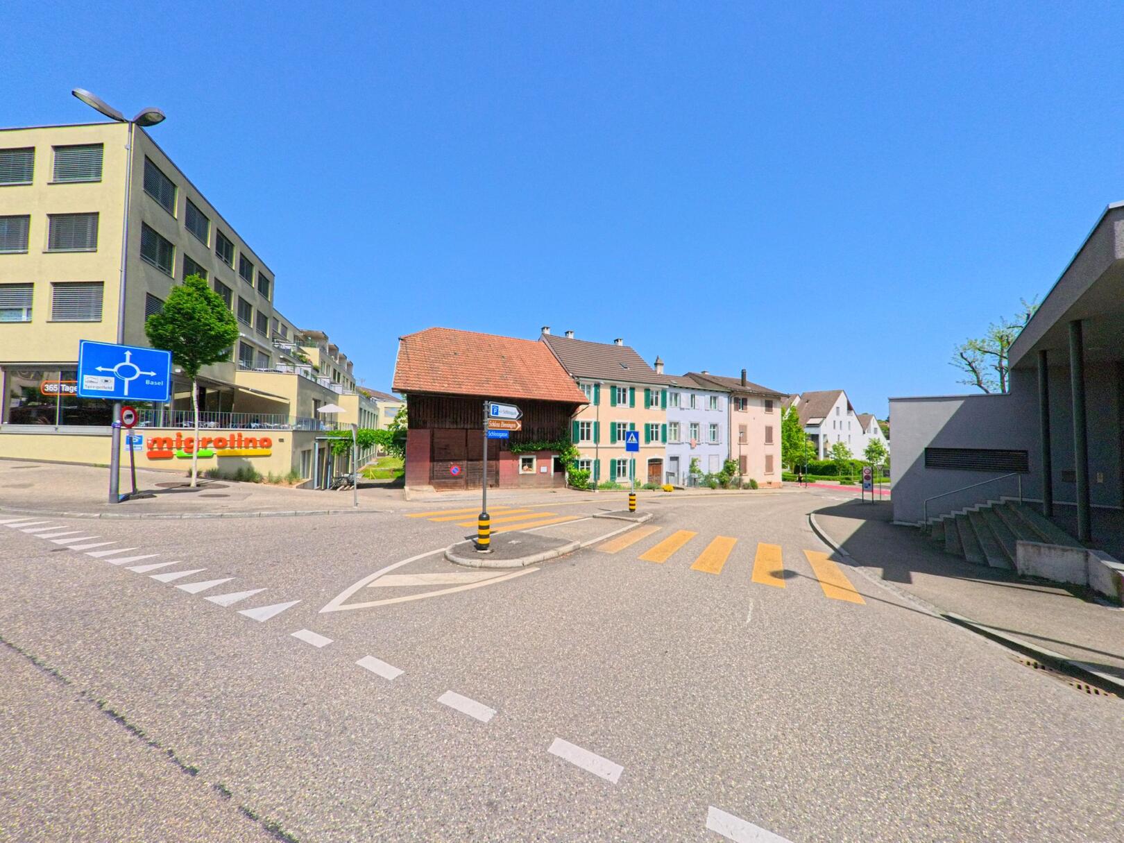 An einer Kreuzung in Binningen (4102, Kanton Basel-Landschaft) stehen Wohnhäuser mit farbigen Fassaden neben modernen Gebäuden und einem Geschäft mit Migrolino-Schild.