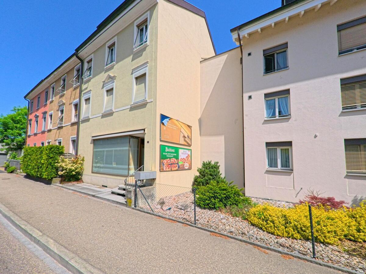 Entlang einer Seitenstrasse in Binningen (4102, Kanton Basel-Landschaft) stehen farbig gestrichene Wohnhäuser mit kleinen Vorgärten und Werbetafeln.
