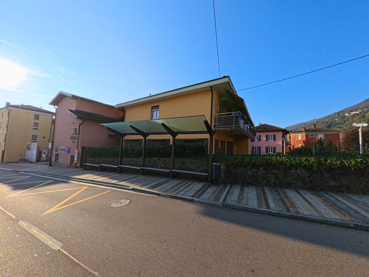An einer Nebenstrasse mit überdachter Bushaltestelle und Wohnhäusern zeigt sich ein ruhiger Abschnitt von Bioggio (6935, Kanton Tessin) bei sonnigem Wetter.