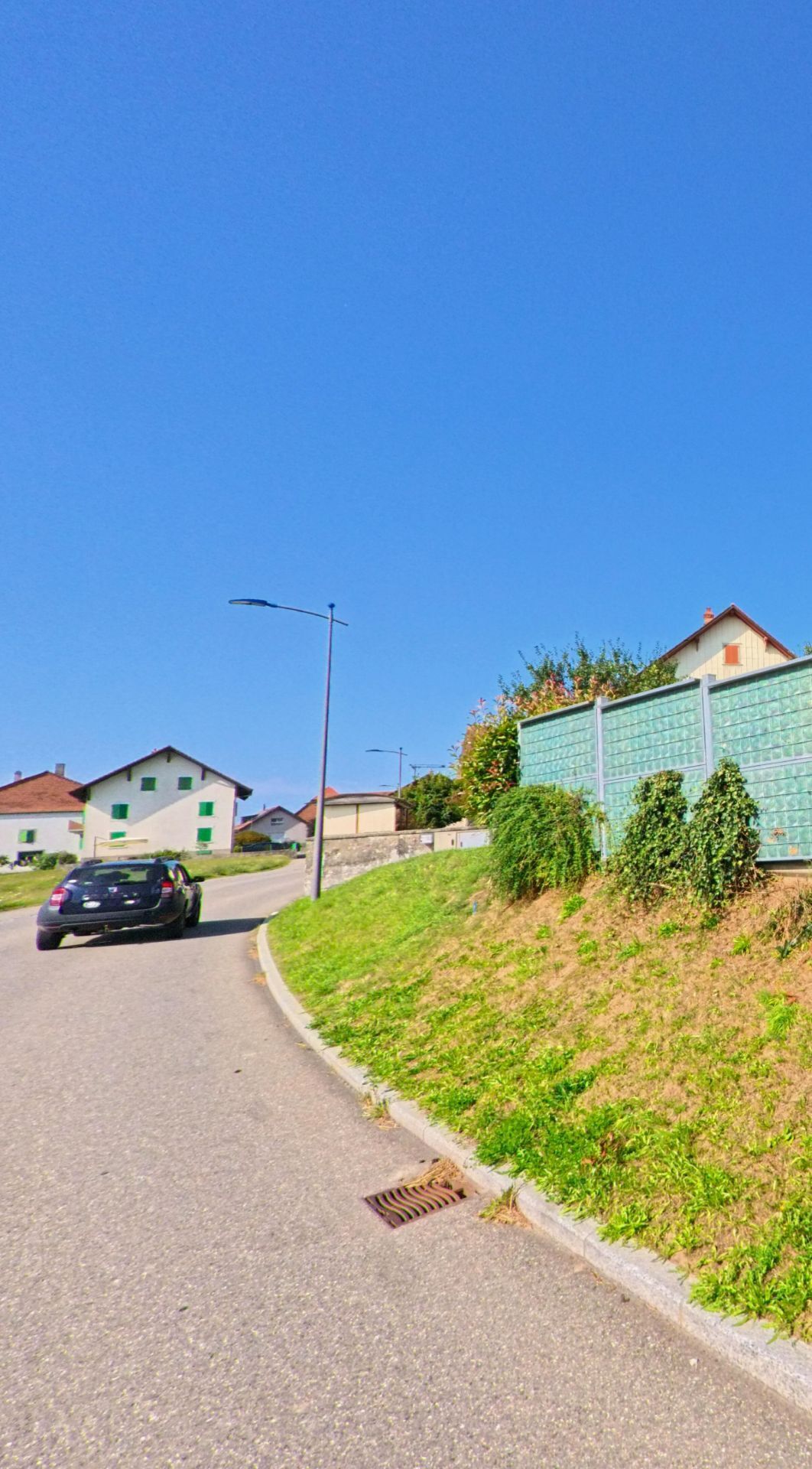 Eine leicht ansteigende Strasse in Bioley-Magnoux (1407, Kanton Waadt) führt an modernen Lärmschutzwänden und Wohnhäusern mit grünen Fensterläden vorbei.