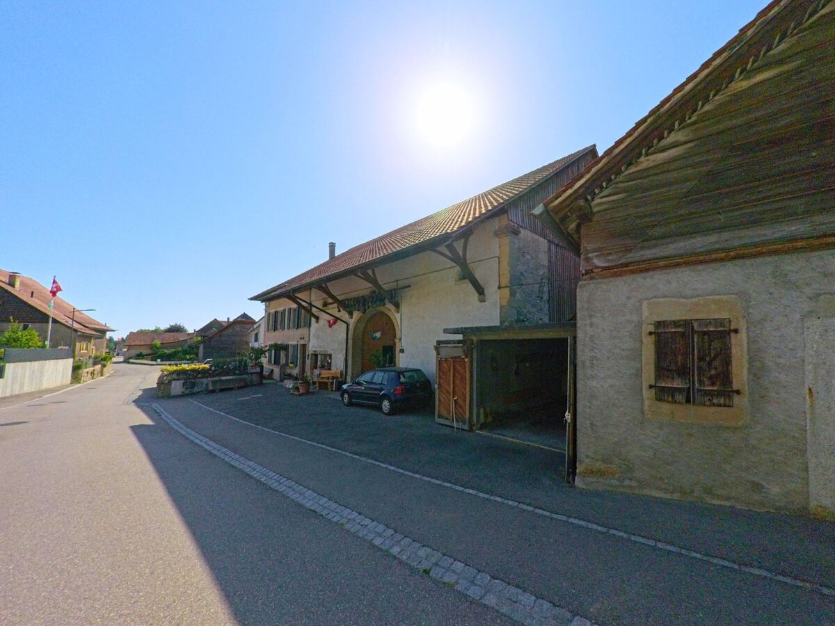 In Bioley-Magnoux (1407, Kanton Waadt) stehen traditionelle Bauernhäuser mit Blumenschmuck und Schweizer Fahnen entlang einer sonnigen Dorfstrasse.