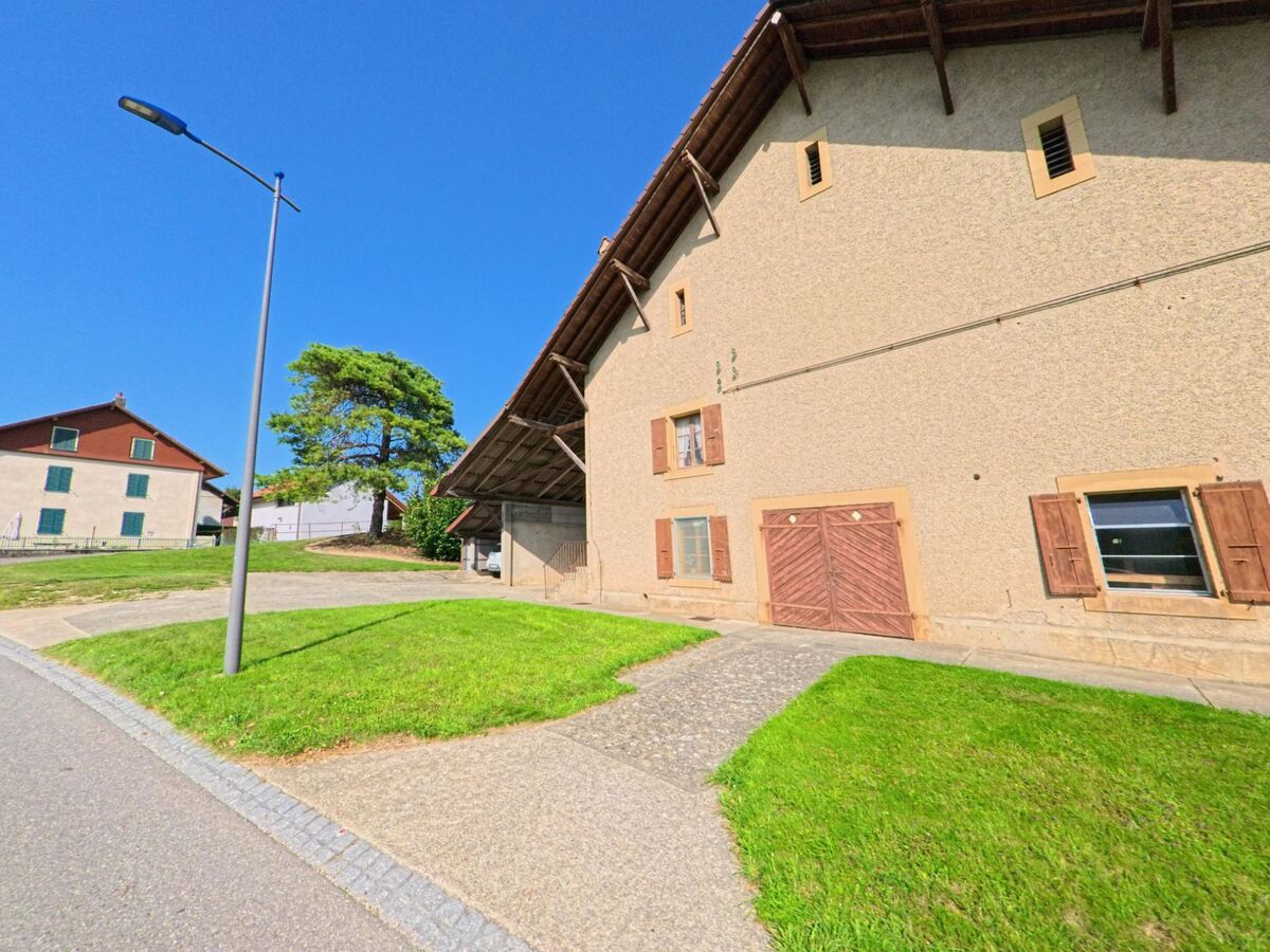 Ein traditionelles Bauernhaus in Bioley-Magnoux (1407, Kanton Waadt) mit Holzläden und gepflegtem Rasen steht an einer leicht ansteigenden Strasse.