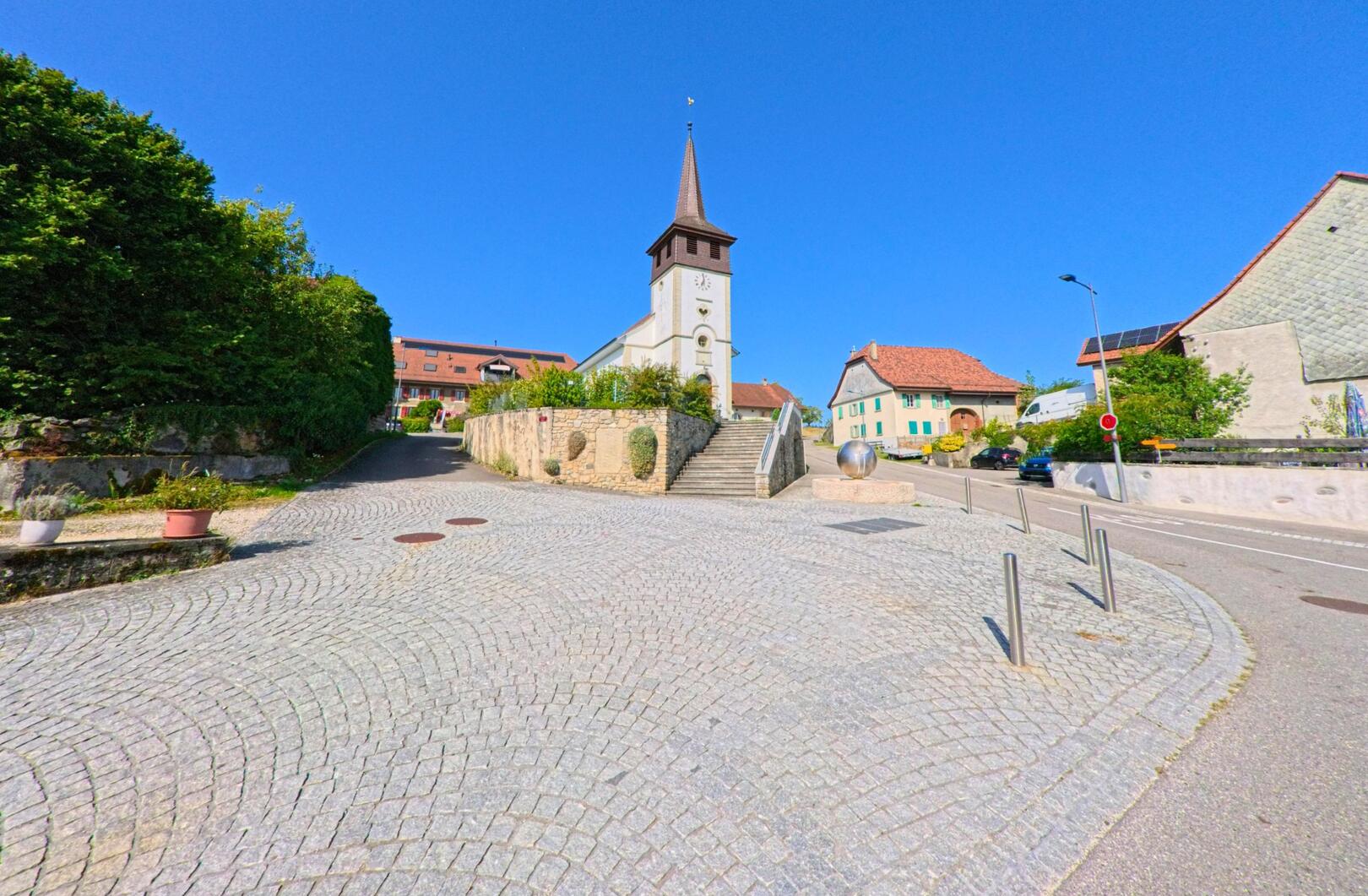 Vor der Kirche von Bioley-Magnoux (1407, Kanton Waadt) liegt ein gepflasterter Platz, umgeben von traditionellen Dorfhäusern unter blauem Himmel.