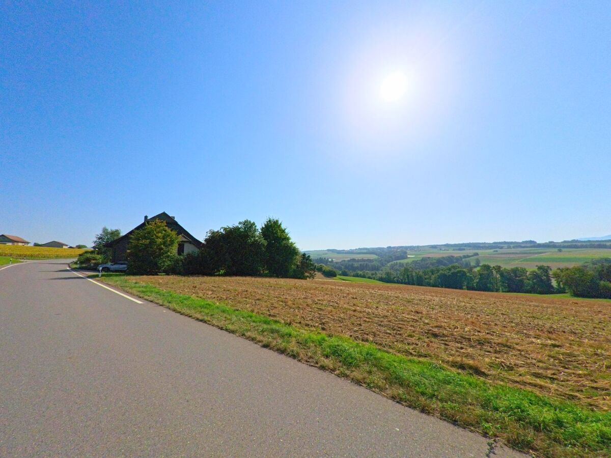 Eine Landstrasse in Bioley-Magnoux (1407, Kanton Waadt) führt an Feldern und einem einzelnen Bauernhaus vorbei mit Blick auf das weite Tal.