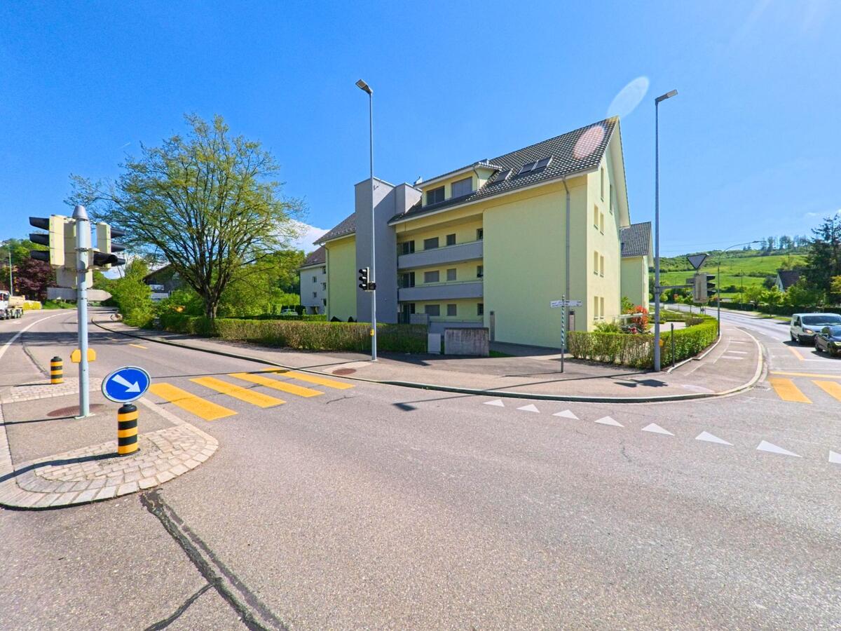 Ein moderner Wohnblock mit heller Fassade steht in Birmensdorf, PLZ 8903 im Kanton Zürich, an einer gut ausgebauten Kreuzung mit Fussgängerstreifen und Verkehrsinseln.