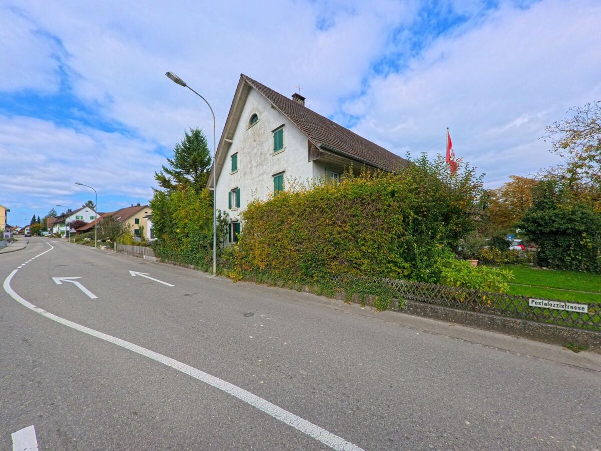 Ein traditionelles Wohnhaus mit gruener Fensterlaeden steht an der Pestalozzistrasse in Birr (5242, Kanton Aargau) und ist von dichter Vegetation umgeben.