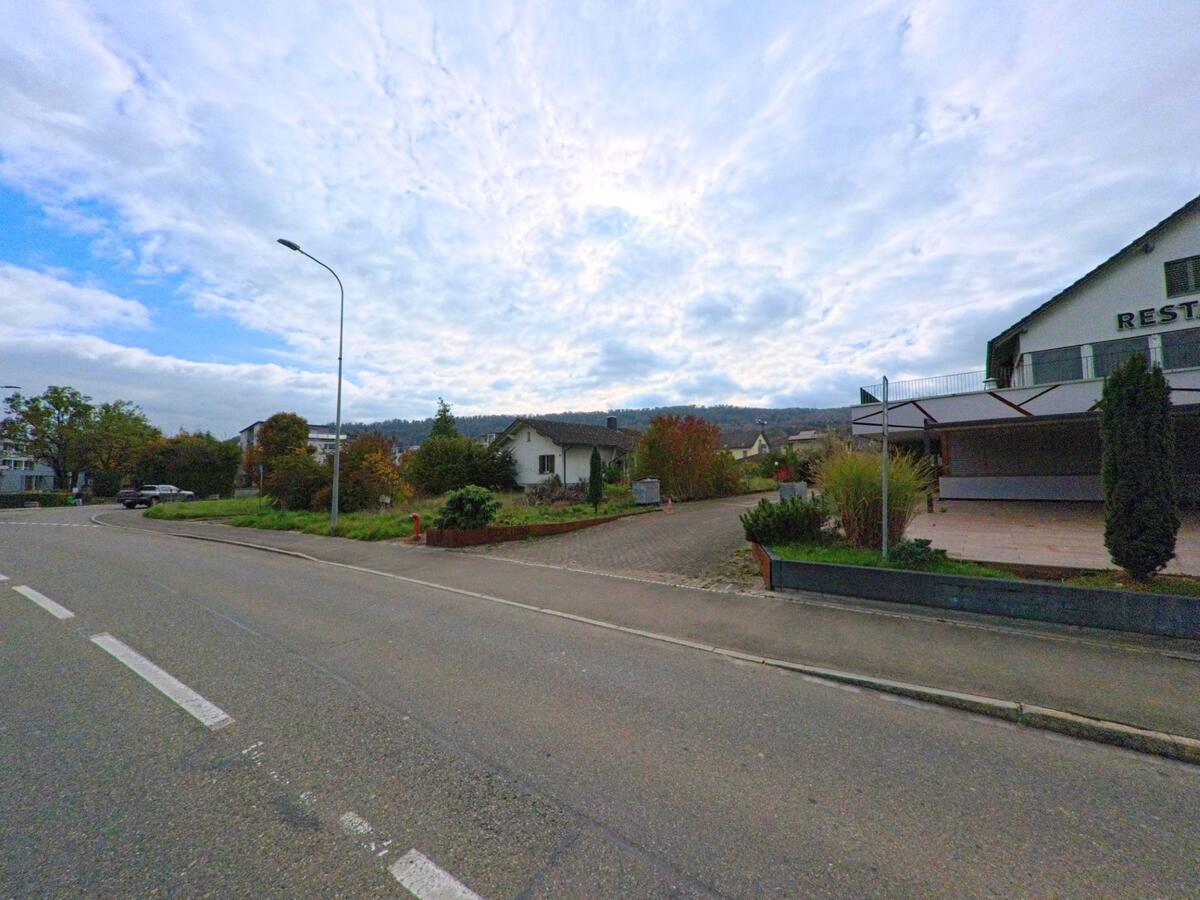 An einer leicht gekruemmten Strasse in Birr (5242, Kanton Aargau) liegen Wohnhaeuser und ein Restaurant mit Terrasse vor bewaldeten Huegeln im Hintergrund.