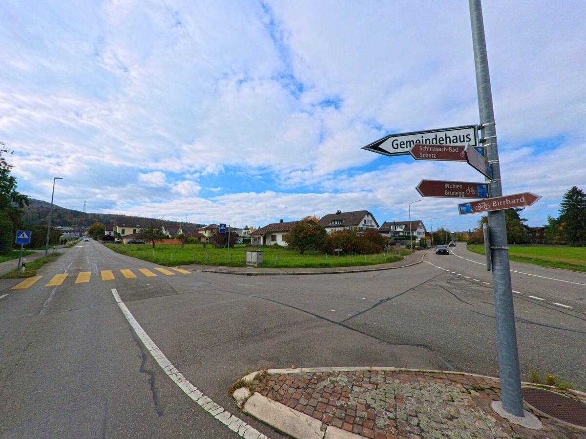An einer Kreuzung in Birr (5242, Kanton Aargau) weisen Wegweiser zu umliegenden Orten wie Scherz, Birrhard und dem Gemeindehaus, waehrend die Strasse durch ein Wohngebiet fuehrt.