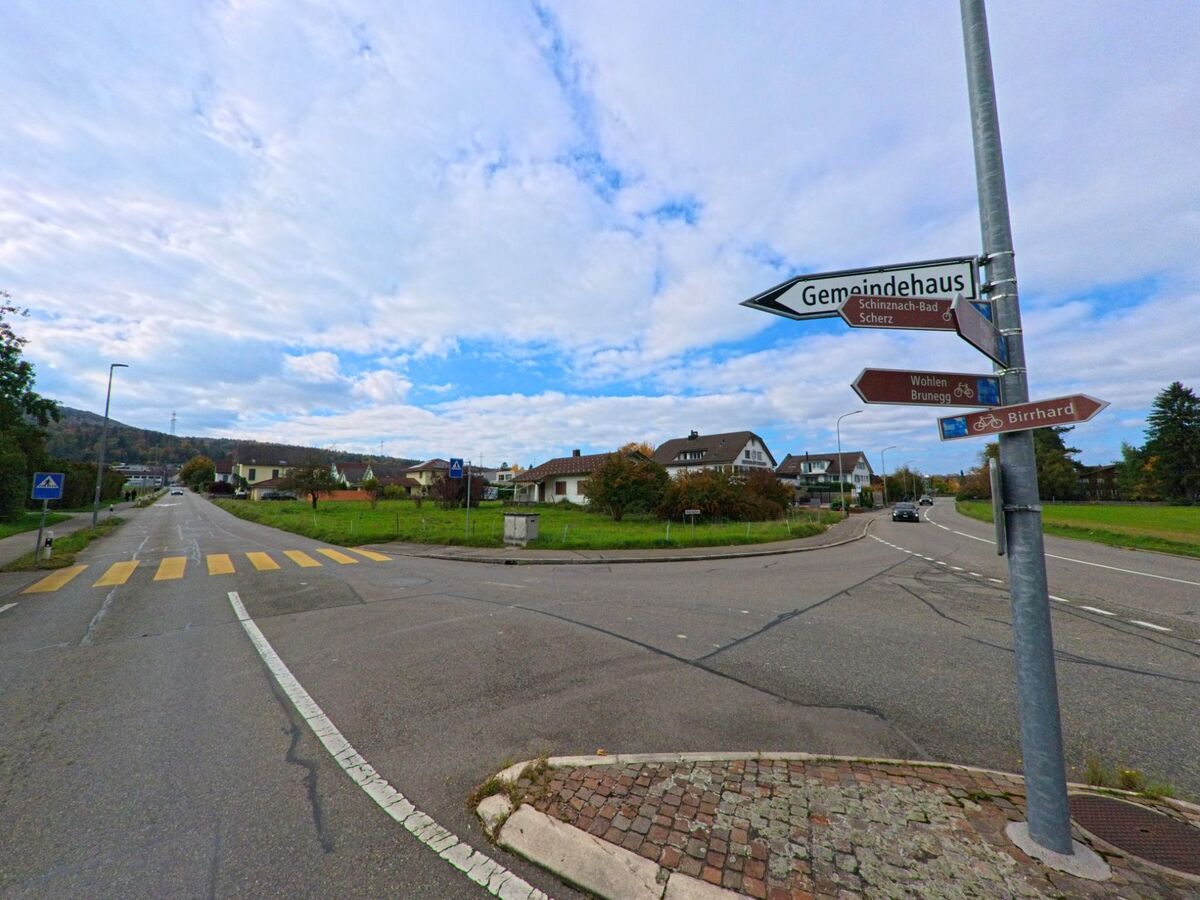 An einer Kreuzung in Birr (5242, Kanton Aargau) weisen Wegweiser zu umliegenden Orten wie Scherz, Birrhard und dem Gemeindehaus, waehrend die Strasse durch ein Wohngebiet fuehrt.