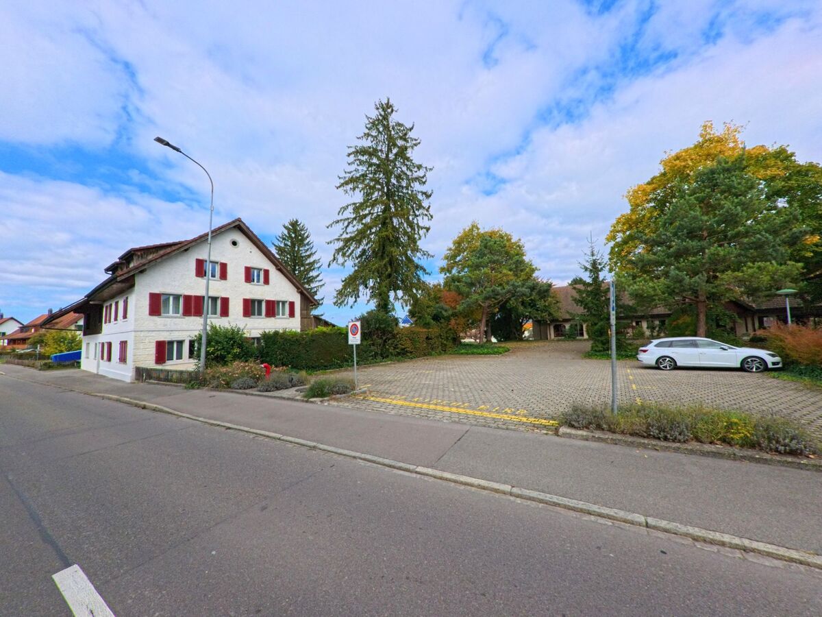 Ein traditionelles Wohnhaus mit roten Fensterlaeden steht in Birr (5242, Kanton Aargau) neben einem grossen Parkplatz mit Baeumen und gepflegter Bepflanzung.