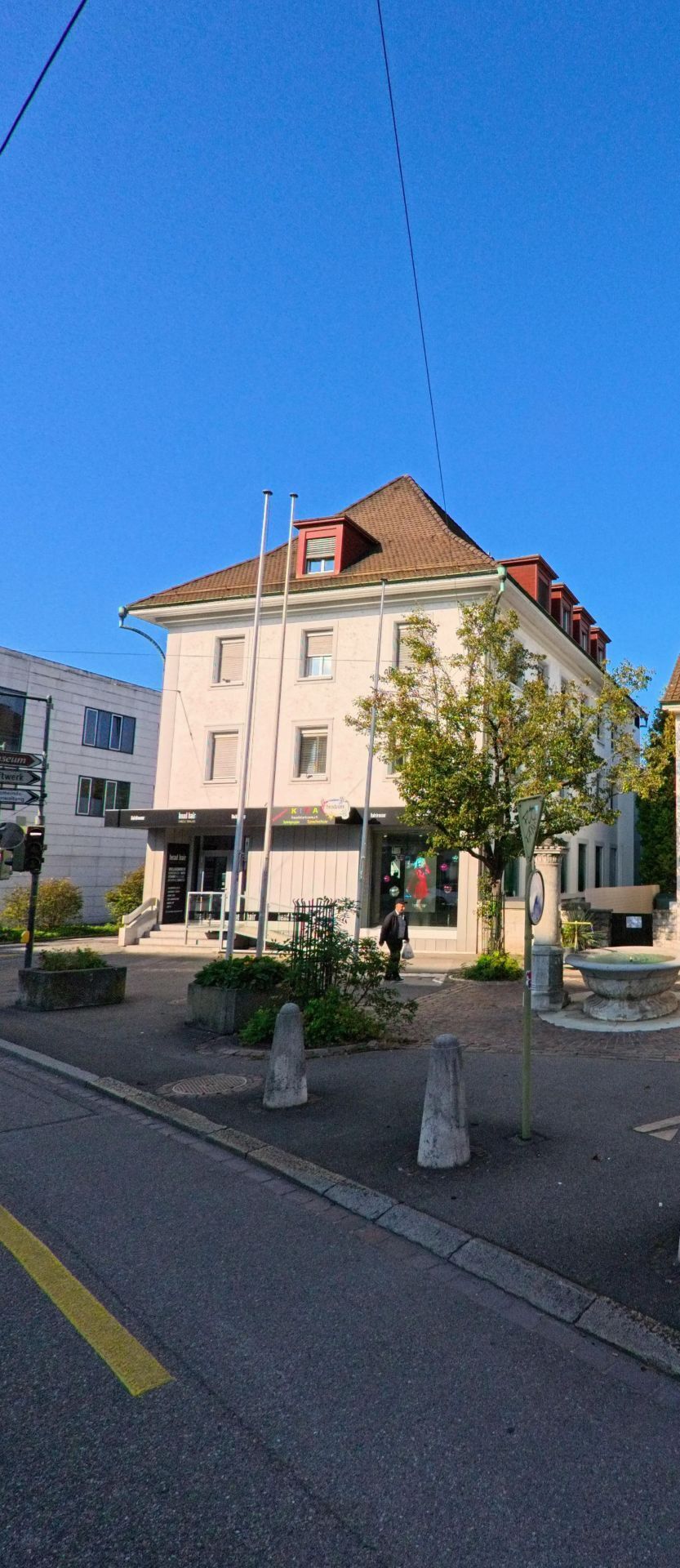 Ein heller Eckbau mit steilem Dach praegt in Birsfelden (4127, Kanton Basel-Landschaft) eine kleine Platzsituation mit Brunnen, Verkehrsinseln und Fussgaengern.