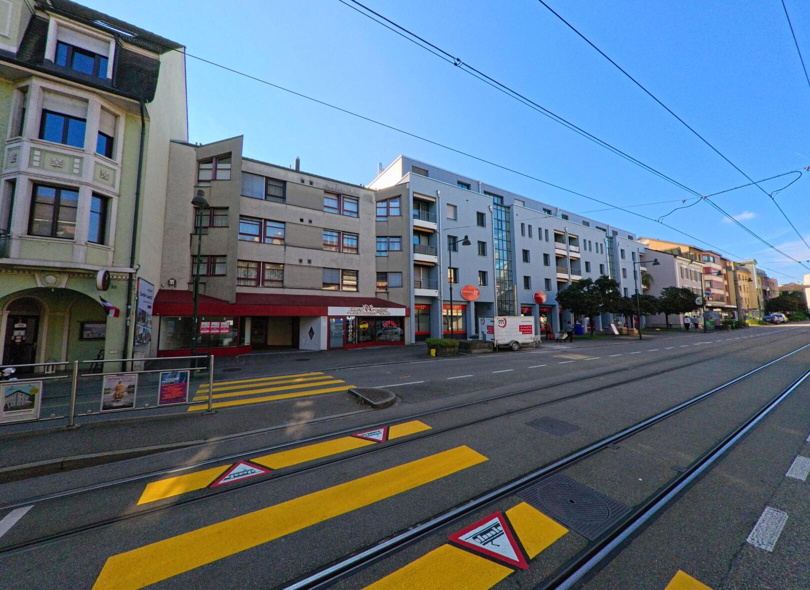 Entlang der belebten Hauptstrasse in Birsfelden (4127, Kanton Basel-Landschaft) stehen mehrgeschossige Wohn- und Geschaeftsgebaeude neben den Tramgleisen im Sonnenlicht.
