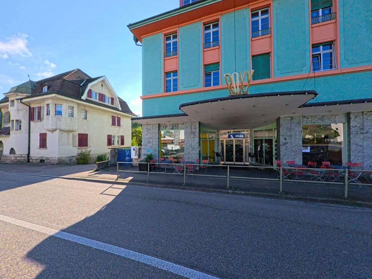 Vor dem markanten Roxy-Gebaeude in Birsfelden (4127, Kanton Basel-Landschaft) stehen rote Sitzgelegenheiten, waehrend daneben ein aelteres Wohnhaus die Strassenszene praegt.