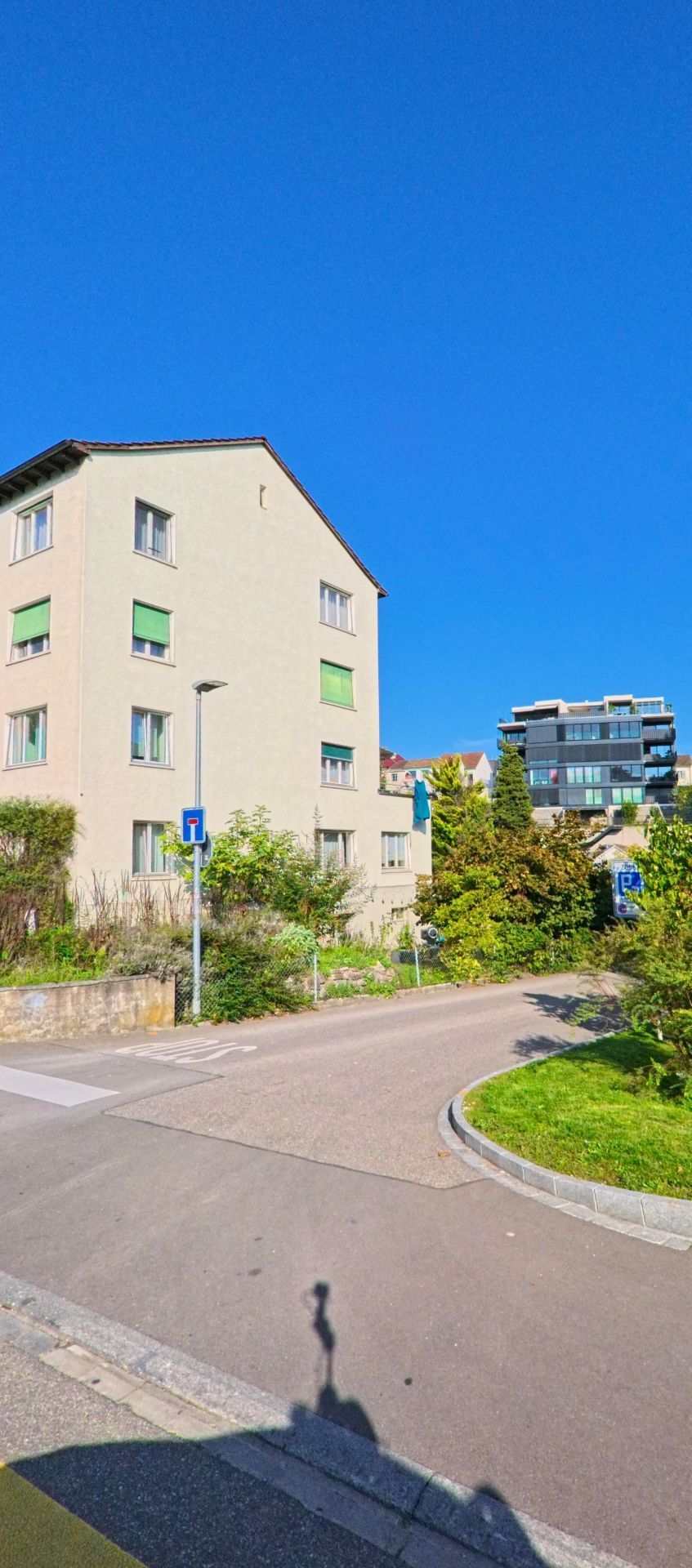 Neben einer kleinen Sackgasse in Birsfelden (4127, Kanton Basel-Landschaft) steht ein schlichtes Wohnhaus mit gruener Umgebung und modernen Gebaeuden im Hintergrund.