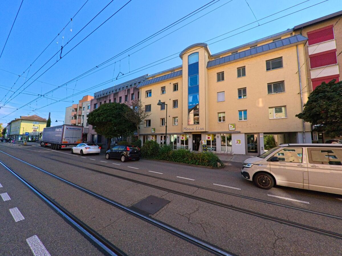 Entlang der Tramlinie in Birsfelden (4127, Kanton Basel-Landschaft) stehen mehrgeschossige Wohn- und Geschaeftsgebaeude, vor denen Fahrzeuge parken und Fussgaenger vorbeigehen.