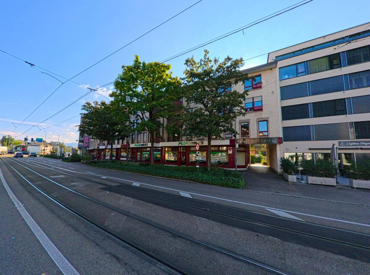 Entlang der Tramtrasse in Birsfelden (4127, Kanton Basel-Landschaft) stehen Laeden und Wohnungen hinter einer Baumreihe, waehrend der Verkehr zur Kreuzung weiterfuehrt.