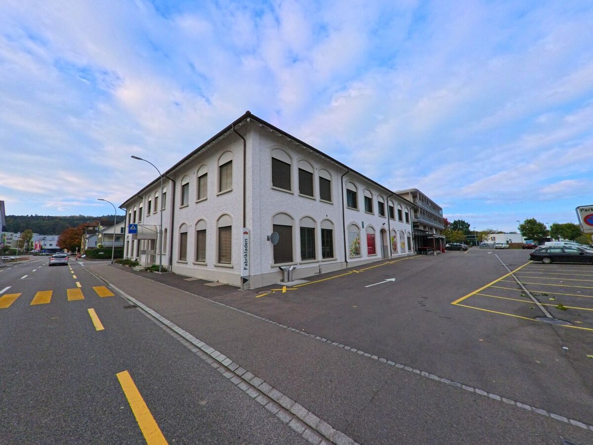 Am Rand eines Parkplatzes in Bischofszell (9220, Kanton Thurgau) steht ein langgestrecktes Gebäude mit Rundbogenfenstern entlang einer ruhig geführten Strasse.