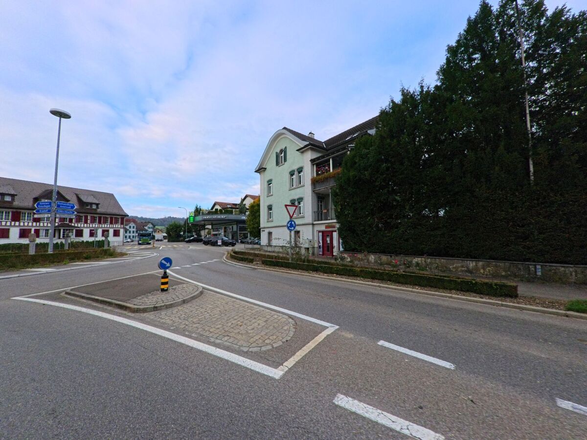 An einem kleinen Verkehrsteiler in Bischofszell (9220, Kanton Thurgau) verlaufen mehrere Strassen zwischen Wohnhaeusern, Hecken und Verkehrszeichen unter hellem Himmel.