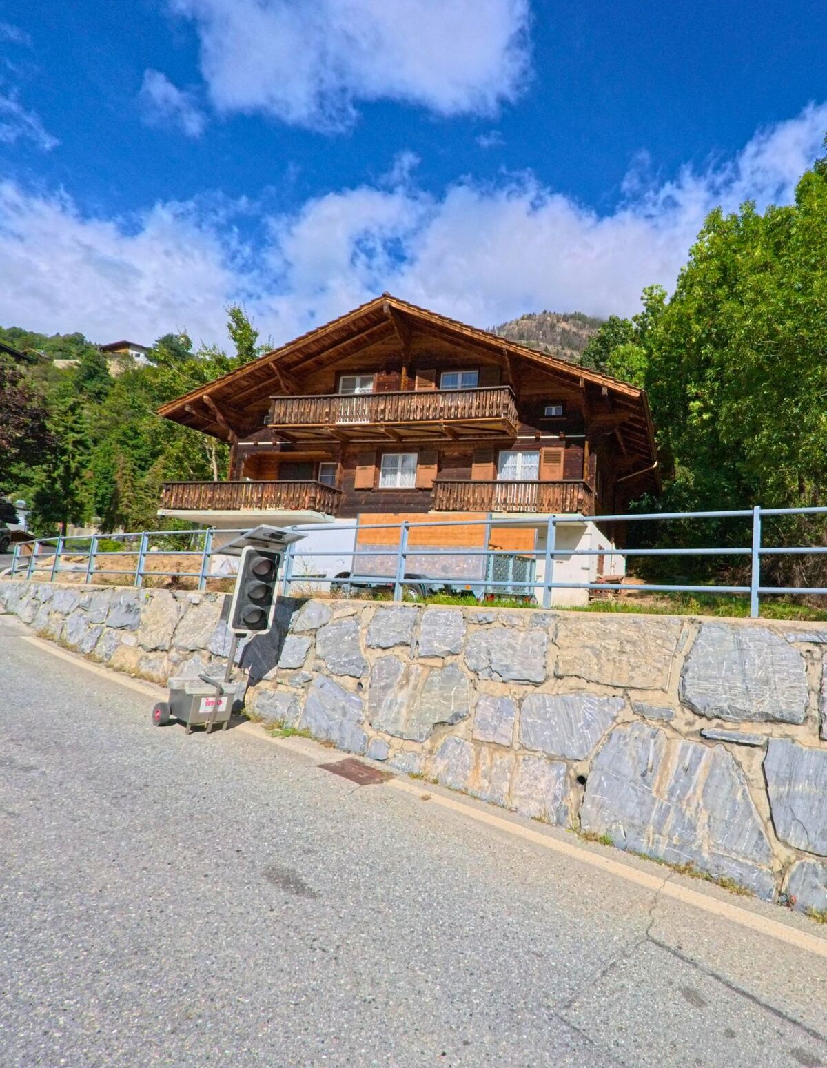 Ein traditionelles Chalet mit Holzfassade und Balkonen erhebt sich in Bitsch (3982, Kanton Wallis) hinter einer Steinmauer an einer ansteigenden Strasse.