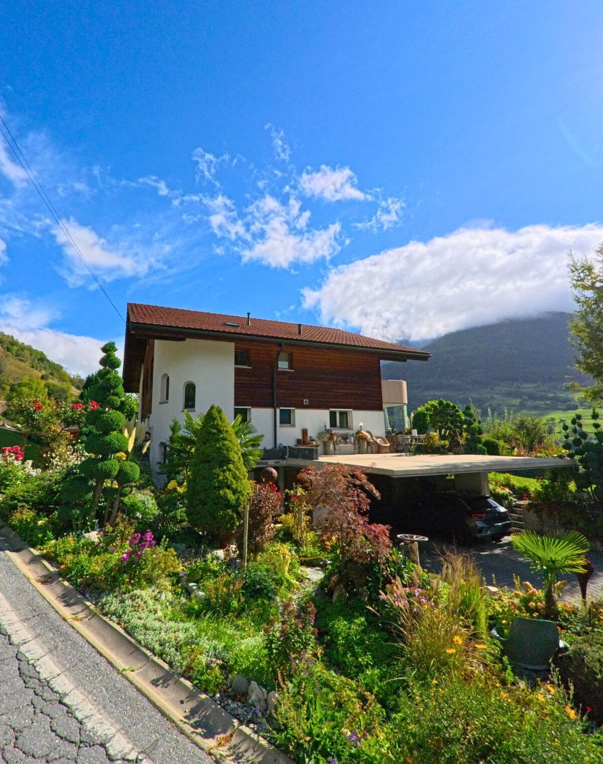 Ein Einfamilienhaus mit Holzfassade in Bitsch (3982, Kanton Wallis) ist von einem bunt bepflanzten Garten umgeben und liegt vor einem bewaldeten Berghang.
