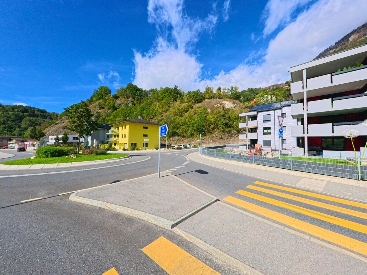 Ein Fussgaengerstreifen fuehrt in Bitsch (3982, Kanton Wallis) ueber die Strasse nahe einem Kreisel mit modernen Wohnhaeusern und einem gelben Gebaeude.