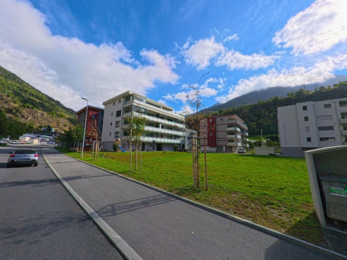 Mehrgeschossige Wohngebaeude in Bitsch (3982, Kanton Wallis) stehen vor einer Gruenflaeche mit jungen Baeumen und den bewaldeten Bergen im Hintergrund.