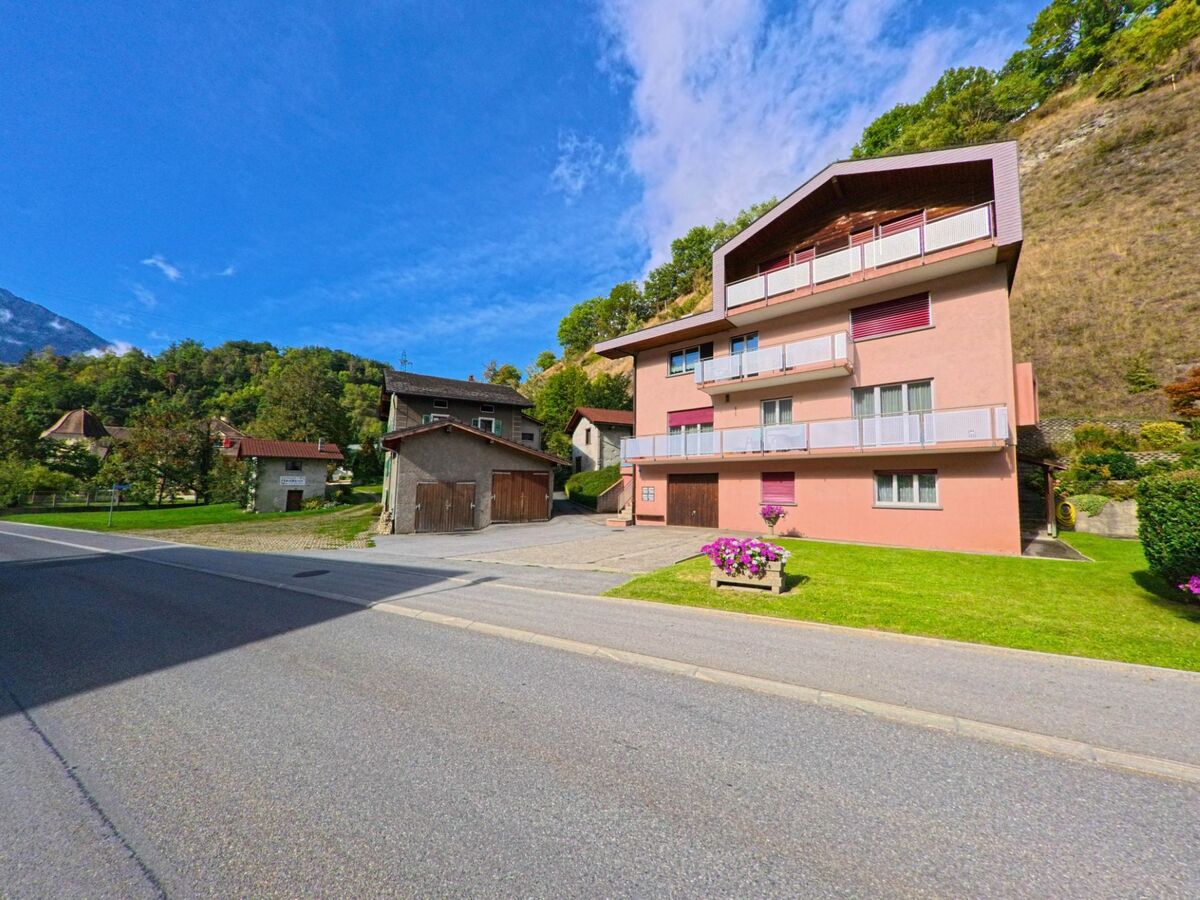 Ein rosa Wohnhaus mit Balkonen steht in Bitsch (3982, Kanton Wallis) am Strassenrand, umgeben von kleineren Gebaeuden und Gruenflaechen.
