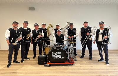 Die Band BlackBoxMusic steht in Reinach (4153, Kanton Baselland) mit Blasinstrumenten und Schlagzeug in einem Proberaum.