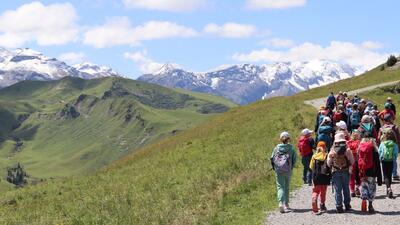 Eine Gruppe von Kindern wandert auf einem Bergweg durch offene Landschaft in Oberwil (4104, Kanton Baselland) mit Blick auf grüne Hügel und schneebedeckte Gipfel.