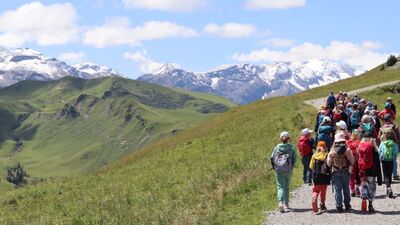 Eine Gruppe von Kindern wandert auf einem Bergweg durch offene Landschaft in Oberwil (4104, Kanton Baselland) mit Blick auf grüne Hügel und schneebedeckte Gipfel.