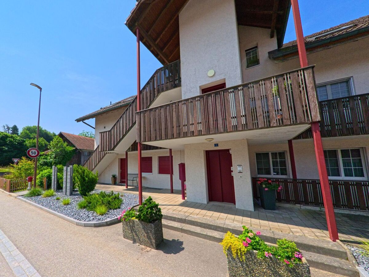 In Bleienbach (3368, Kanton Bern) zeigt sich ein modernes Wohngebäude mit Holzverkleidung, rotem Eingangsbereich und gepflegtem Vorgarten an einer verkehrsberuhigten Strasse.