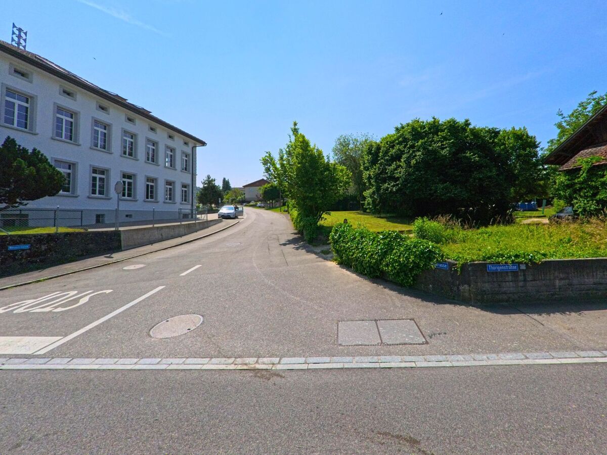 In Bleienbach (3368, Kanton Bern) verläuft die Thörigenstrasse an einem weissen Schulgebäude vorbei in eine leicht ansteigende Wohngegend mit viel Grünfläche.