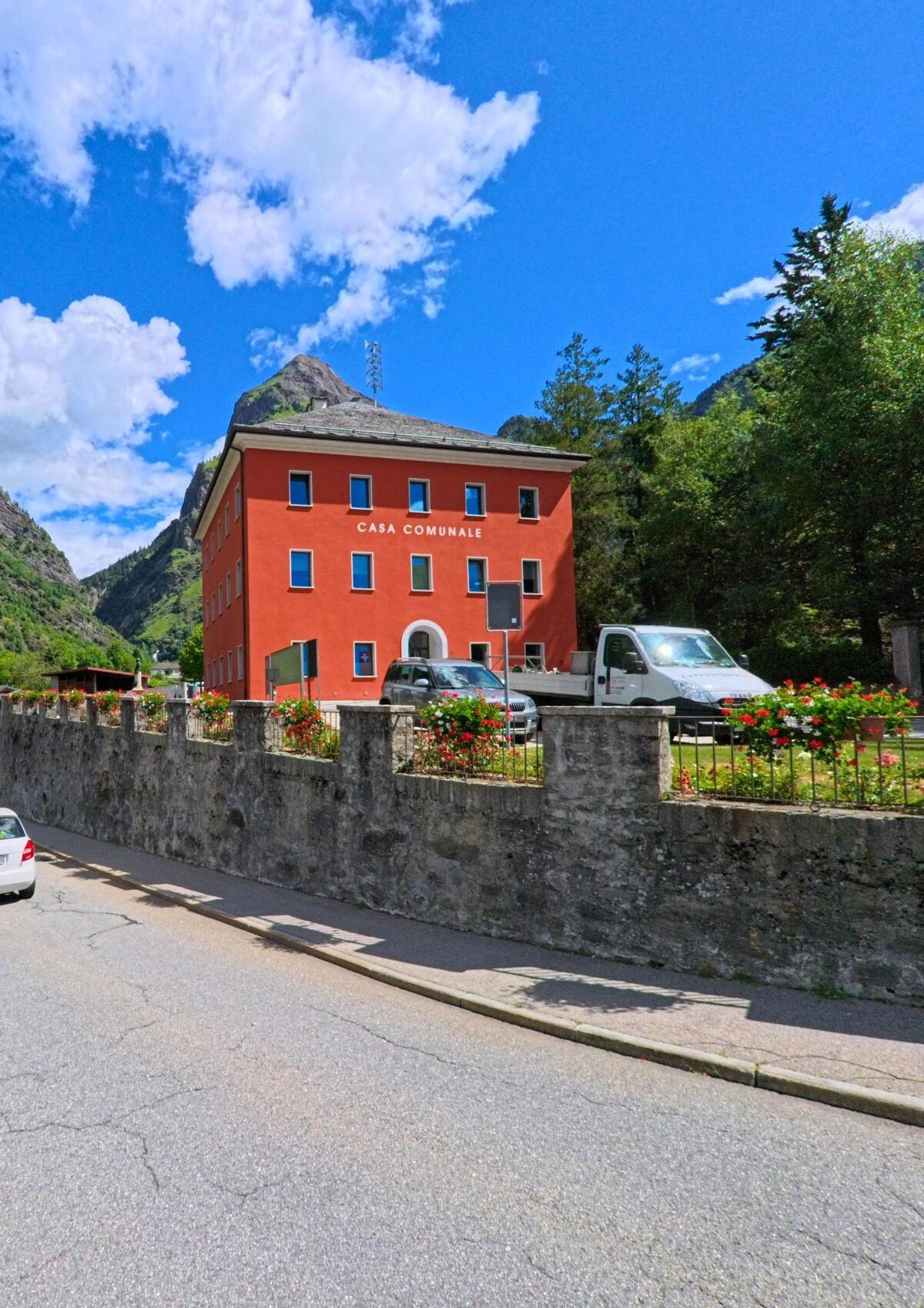Das rote Gemeindehaus mit der Aufschrift „Casa Comunale“ steht in Blenio (6718, Kanton Tessin) vor einer Bergkulisse und ist von Blumenbeeten und einer Steinmauer umgeben.