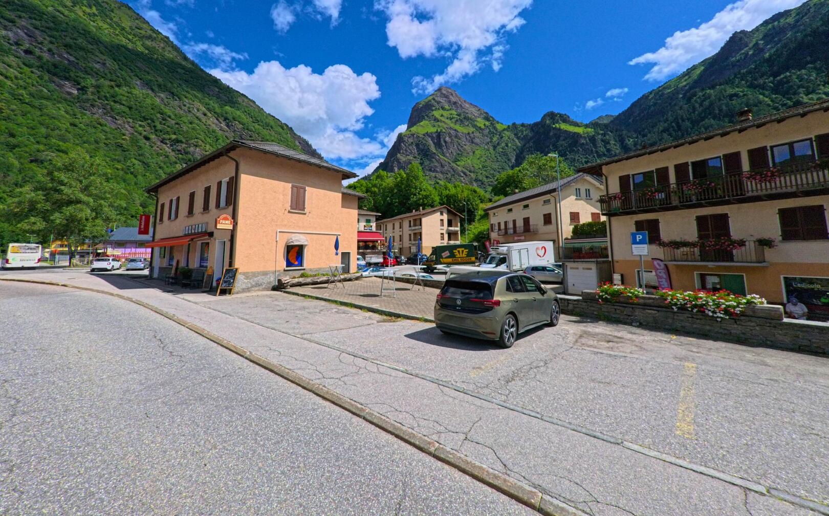 Im Zentrum von Blenio (6718, Kanton Tessin) stehen Wohn- und Geschaeftshaeuser mit Blumenbalkonen und einer kleinen Baeckerei, eingerahmt von steilen Berghaengen.