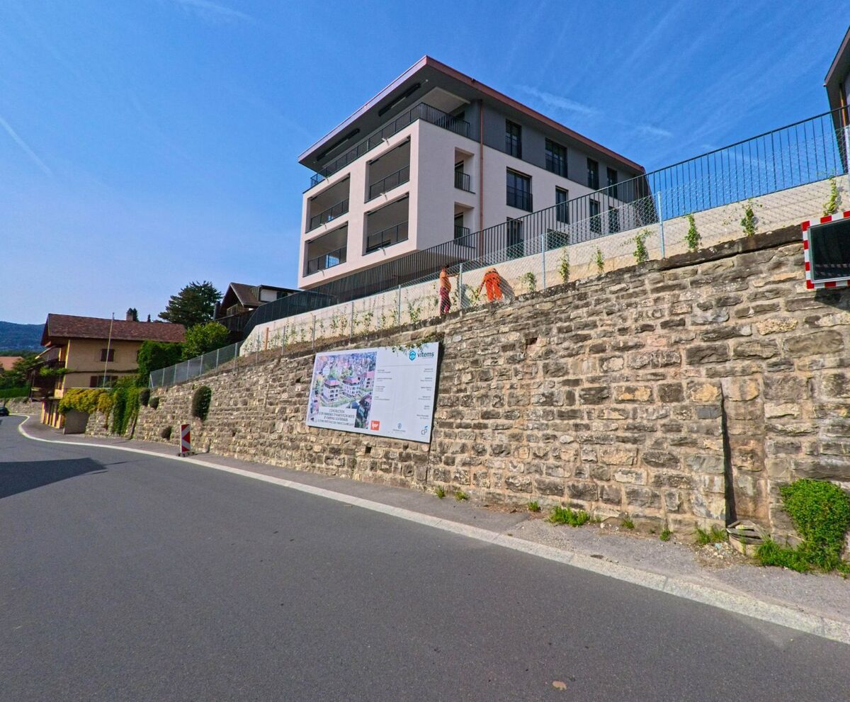 Ein modernes Mehrfamilienhaus erhebt sich in Blonay_-_Saint-Legier (1807, Kanton Waadt) oberhalb einer alten Steinmauer mit Bauhinweisschild.