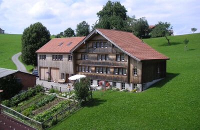 In Waldstatt (9104, Kanton Appenzell Ausserrhoden) liegt das BnB Frischknecht in einem traditionellen Holzhaus mit rotem Ziegeldach, umgeben von gruenen Wiesen und Gartenanlage.