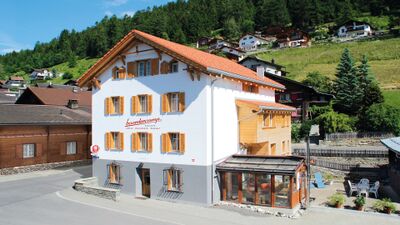 In Ruschein (7154, Kanton Graubuenden) steht das Boardercamp Swiss Mountain Hostel mit weisser Fassade, Holzlaeden und einem kleinen Aussenbereich.