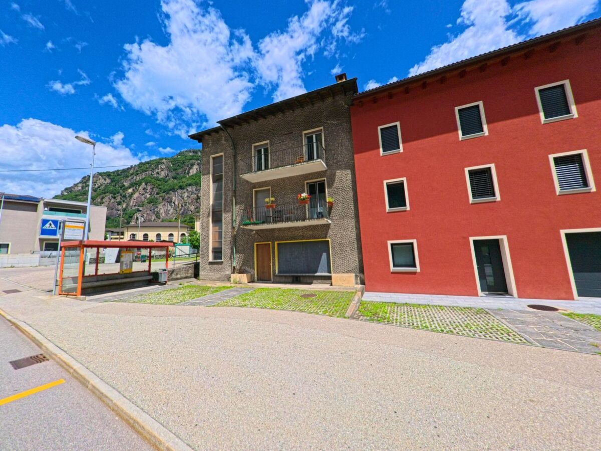 In Bodio (6743, Kanton Tessin) steht eine kleine Bushaltestelle vor zwei nebeneinanderliegenden Wohnhaeusern mit roten und dunklen Fassaden am Fuss der Berge.