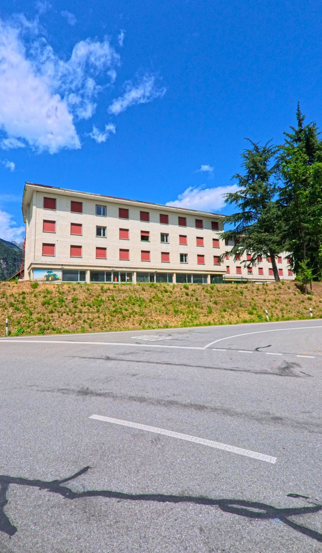 Ein helles Gebaeude mit roten Fensterlaeden steht in Bodio (6743, Kanton Tessin) oberhalb einer Strasse, neben hohen Baeumen und unter einem klaren blauen Himmel.